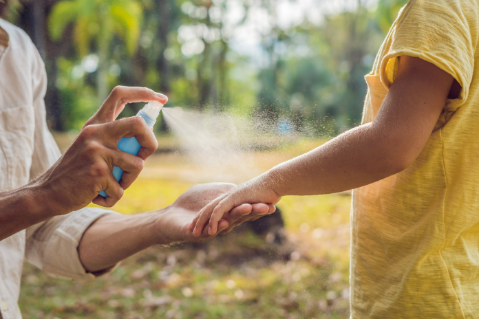 Mother and child are outside. Mother is spraying mosquito repellent on her child's arm.